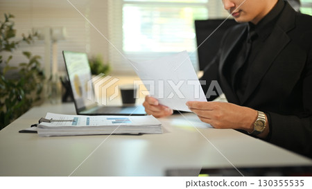 Businessman in formal suit reviewing financial documents at modern office desk 130355535