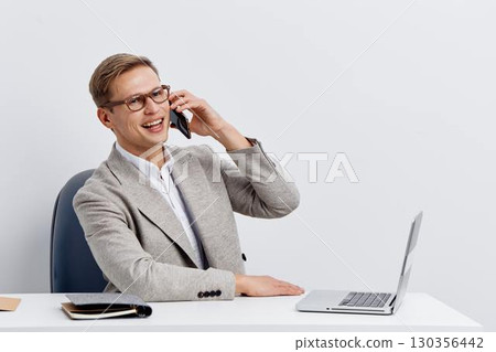 Professional smiling man in formal attire talking on mobile phone while seated at desk with laptop isolated on plain white background studio portrait. Business workplace concept Professional smiling man in formal attire talking on mobile phone while seated at desk with laptop isolated on plain white background studio portrait. Business workplace concept 130356442