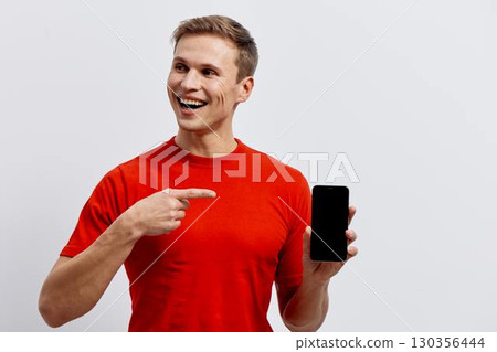 smiling young caucasian man wear red t-shirt hold smartphone black screen point finger isolated on plain gray background digital nomad technology concept smiling young caucasian man wear red t-shirt hold smartphone black screen point finger isolated on plain gray background digital nomad technology concept 130356444