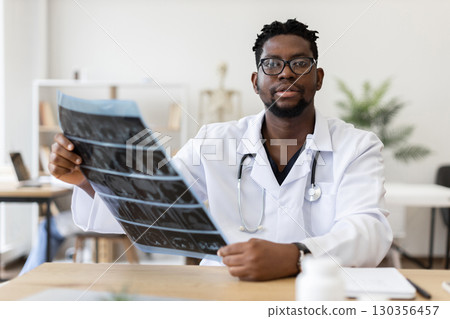 Male African doctor wearing white coat with stethoscope analyzing X-ray in modern office during daytime, demonstrating care for patient health and diagnosis processing 130356457