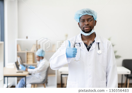 Male doctor in medical attire with stethoscope giving thumbs-up gesture, medical office background. Confident gesture represents healthcare professional optimism and support. 130356483