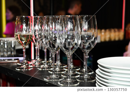 Empty wine glasses neatly arranged on a bar counter with a stack of plates, prepared for a night party and festive celebration atmosphere. Empty wine glasses neatly arranged on a bar counter with a stack of plates, prepared for a night party and festive celebration atmosphere. 130357750