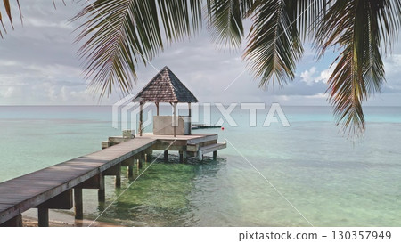 French Polynesia, Rangiroa island: Palm tree leaves framing wooden pier and gazebo over turquoise lagoon coastal area, clear waters reflect the sky, creating a peaceful atmosphere ideal for relaxation 130357949