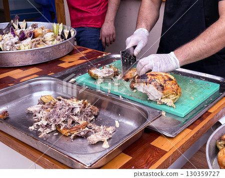 Cook slicing roasted chicken on a cutting board in a kitchen setting. Culinary preparation, food service, and gastronomy. 130359727