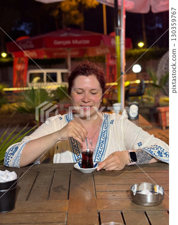 Woman stirring Turkish tea in a traditional glass at a cafe table. Evening leisure, cultural travel, and authentic lifestyle during vacation. 130359767