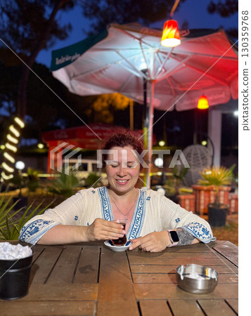 Woman sitting at an outdoor cafe table with a glass of Turkish tea. Leisure, travel, and authentic cultural experience during evening relaxation. Woman sitting at an outdoor cafe table with a glass of Turkish tea. Leisure, travel, and authentic cultural experience during evening relaxation. 130359768