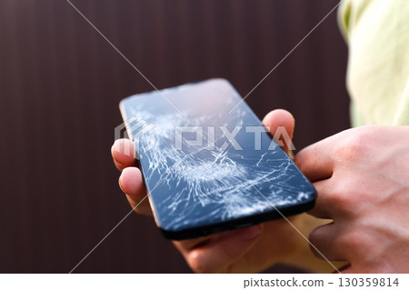 a male hand holding a phone with a broken screen. broken phone screen repair. male hand holding shattered smartphone. mobile device damage problem a male hand holding a phone with a broken screen. broken phone screen repair. male hand holding shattered smartphone. mobile device damage problem 130359814