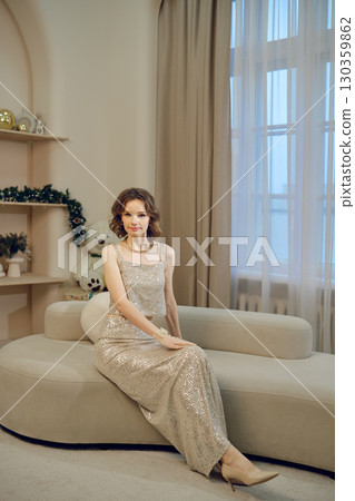 Elegant woman in a sparkling dress seated on a stylish sofa in a cozy indoor setting Elegant woman in a sparkling dress seated on a stylish sofa in a cozy indoor setting 130359862
