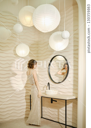 Elegant woman in a shimmering dress stands near a stylish bathroom mirror Elegant woman in a shimmering dress stands near a stylish bathroom mirror 130359863