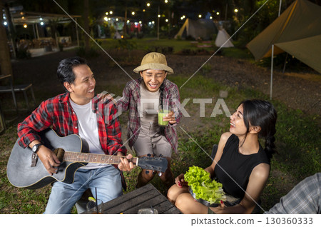 Indonesian southeast asian people cooking sliced marinated beef bulgogi on a grill pan. One of them is holding an acoustic guitar. Scene of the lifestyle of enjoying food in a natural setting 130360333