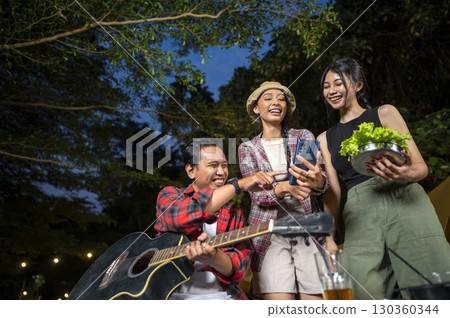 Indonesian southeast asian people enjoying quality time together. Man holds a guitar, and the other women show something on her cellphone. Scene of a lifestyle of enjoying camping in a natural setting Indonesian southeast asian people enjoying quality time together. Man holds a guitar, and the other women show something on her cellphone. Scene of a lifestyle of enjoying camping in a natural setting 130360344