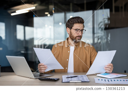 A professional man wearing glasses and a casual shirt examines documents in an office setting. A laptop and charts are on the desk. The environment suggests a modern and dynamic workplace. A professional man wearing glasses and a casual shirt examines documents in an office setting. A laptop and charts are on the desk. The environment suggests a modern and dynamic workplace. 130361213