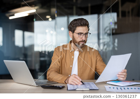 Focused businessman wearing eyeglasses analyzing financial documents and using laptop while sitting at desk in modern office, working with graphs and charts Focused businessman wearing eyeglasses analyzing financial documents and using laptop while sitting at desk in modern office, working with graphs and charts 130361237