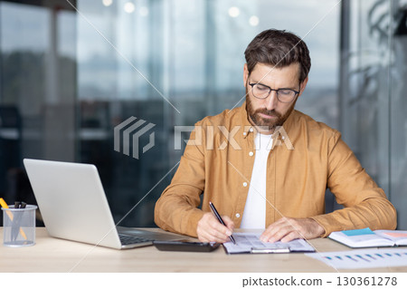 A focused businessman in glasses reviews documents at his desk, highlighting a concept of financial analysis. A focused businessman in glasses reviews documents at his desk, highlighting a concept of financial analysis. 130361278