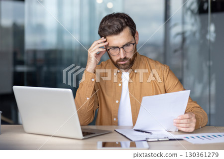 Stressed businessman reading through documents and rubbing his temples while working on a laptop at an office desk, grappling with overwhelming paperwork and mounting pressure 130361279