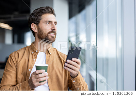 A man with a beard holds a phone and a coffee cup, looking out the window in an office setting. 130361306