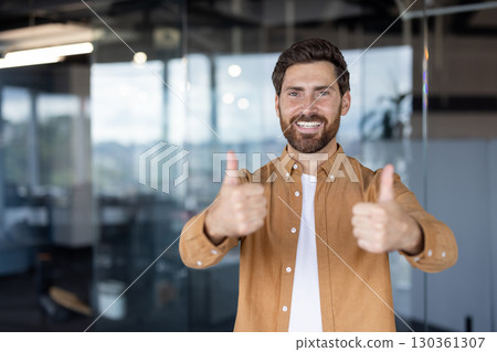 Smiling man with beard showcasing positivity and confidence by giving two thumbs up in a bright and professional office environment. Smiling man with beard showcasing positivity and confidence by giving two thumbs up in a bright and professional office environment. 130361307