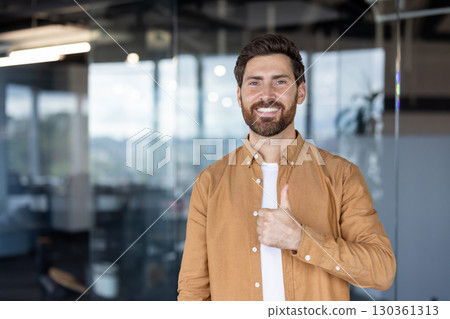 A smiling, bearded man gives a thumbs-up in a modern office setting, exuding positivity and approval. A smiling, bearded man gives a thumbs-up in a modern office setting, exuding positivity and approval. 130361313
