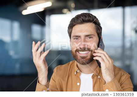 Bearded entrepreneur gesturing while speaking on mobile phone, enjoying positive business conversation in modern office, expressing success and satisfaction 130361359