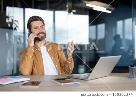 A man in his office, talking on his phone while smiling. He's working on his laptop with a pleasant expression. 130361374