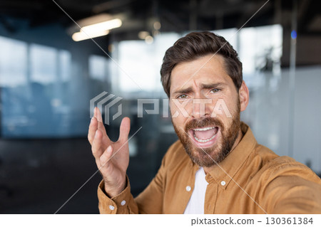 Stressed businessman shouting during a video call in the office, conveying deep frustration and anger stemming from overwhelming work-related challenges and pressures. 130361384