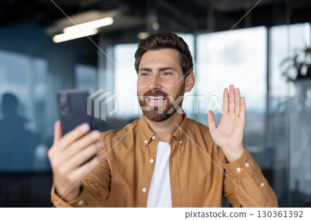 A man waves during a video call on his smartphone inside an office setting, smiling and looking at the screen. A man waves during a video call on his smartphone inside an office setting, smiling and looking at the screen. 130361392