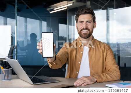 A smiling man in a brown shirt holds up a smartphone with a blank white screen, suitable for displaying an app, surrounded by office elements. 130361417