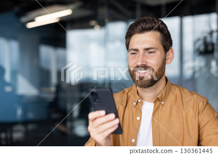 A cheerful man in casual attire is using his smartphone inside a modern office. The contemporary setting includes glass walls and bright lighting, creating a professional yet comfortable atmosphere. A cheerful man in casual attire is using his smartphone inside a modern office. The contemporary setting includes glass walls and bright lighting, creating a professional yet comfortable atmosphere. 130361445