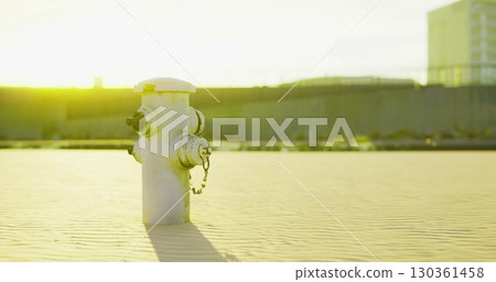 In a bright morning glow, a fire hydrant stands alone on a sandy surface near an urban construction site. In a bright morning glow, a fire hydrant stands alone on a sandy surface near an urban construction site. 130361458