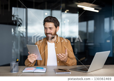 A smiling man in a modern office setting, looking at a tablet during a video call, with a laptop and notebook on the desk. A smiling man in a modern office setting, looking at a tablet during a video call, with a laptop and notebook on the desk. 130361469