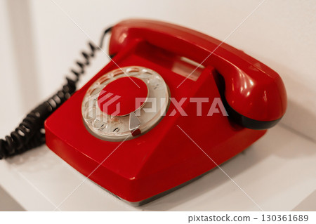 A bright red rotary telephone rests on a white shelf, contrasting with the minimalist decor of the room. Soft lighting highlights its retro design, evoking nostalgia 130361689