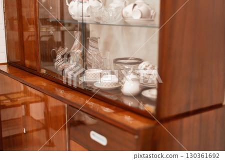 A wooden display cabinet showcases an assortment of vintage dishware, including plates and teacups, arranged neatly on glass shelves. Sunlight illuminates the elegant pieces A wooden display cabinet showcases an assortment of vintage dishware, including plates and teacups, arranged neatly on glass shelves. Sunlight illuminates the elegant pieces 130361692