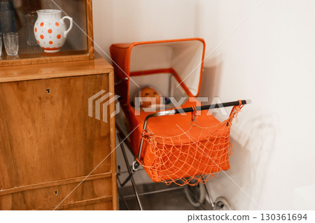 A charming vintage orange baby stroller holds a doll, placed against a wooden cabinet in a cozy indoor space, highlighting nostalgic decor and styling A charming vintage orange baby stroller holds a doll, placed against a wooden cabinet in a cozy indoor space, highlighting nostalgic decor and styling 130361694