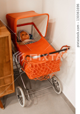 A vintage doll rests comfortably in an orange stroller situated in a warm room corner. The scene evokes nostalgia with its soft lighting and charming decor A vintage doll rests comfortably in an orange stroller situated in a warm room corner. The scene evokes nostalgia with its soft lighting and charming decor 130361696