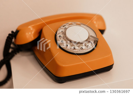 A bright orange rotary telephone is positioned on a white table, highlighting its retro design. This classic phone evokes nostalgia and showcases intricate craftsmanship common in earlier decades 130361697