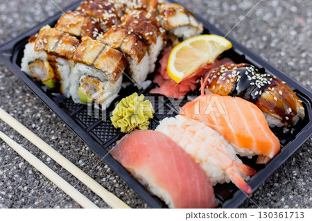 A diverse selection of sushi is arranged on a black tray, showcasing rolls topped with eel and lemon slices alongside vibrant nigiri and wasabi in daylight 130361713