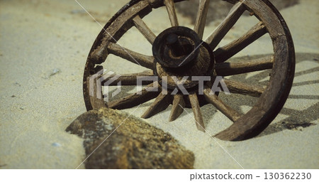 A weathered wooden wagon wheel lies partially buried in sand beside a large stone. The seaside landscape features a gentle slope of sand with hints of a coastal environment. 130362230