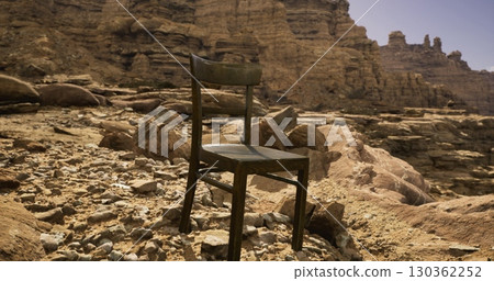 A wooden chair stands isolated on uneven rocks in an arid desert environment. The bright midday sun casts shadows on the rugged terrain, highlighting the stark beauty of the location. A wooden chair stands isolated on uneven rocks in an arid desert environment. The bright midday sun casts shadows on the rugged terrain, highlighting the stark beauty of the location. 130362252