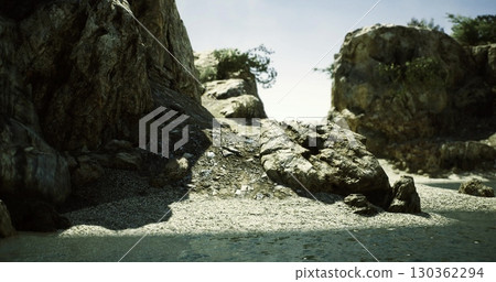 Rocky coastline with textured stones and scattered plant life near tranquil water under a clear blue sky during daylight. Natural setting evokes peace and solitude. 130362294