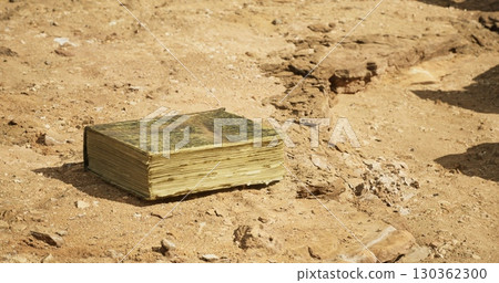 An aged book rests on dry, cracked earth surrounded by rocky terrain. Sunlight illuminates its weathered pages, hinting at stories long forgotten in this desolate location. An aged book rests on dry, cracked earth surrounded by rocky terrain. Sunlight illuminates its weathered pages, hinting at stories long forgotten in this desolate location. 130362300