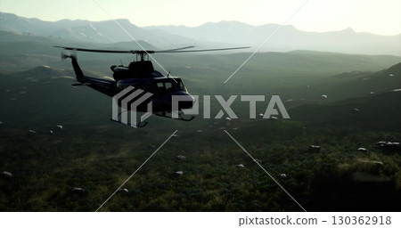 A helicopter hovers gracefully above vast green valleys, surrounded by majestic mountains under a warm sunset sky. The peaceful terrain below contrasts with the machines speed. 130362918
