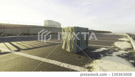 A wrapped object sits quietly on a peaceful beachfront pathway, surrounded by the golden glow of early morning sun and distant high rise buildings. The atmosphere feels tranquil yet intriguing. 130363008