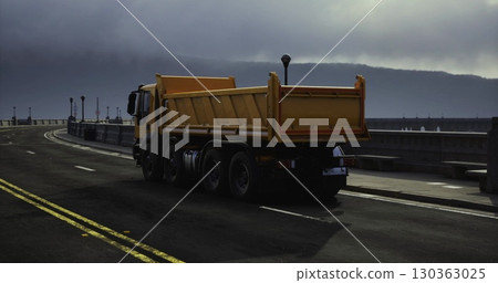 A vibrant yellow dump truck navigates a deserted coastal road. The gloomy clouds hang low in the sky, creating an atmospheric backdrop. Street lamps line the roadway, adding to the serene ambiance. 130363025