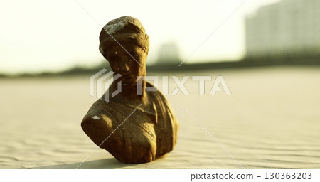 A weathered bronze bust of a woman stands prominently amidst the golden sand, illuminated by soft rays of the setting sun. Gentle waves lap at the shore nearby. A weathered bronze bust of a woman stands prominently amidst the golden sand, illuminated by soft rays of the setting sun. Gentle waves lap at the shore nearby. 130363203