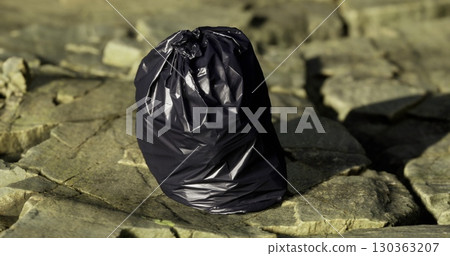 A dark trash bag rests on a rugged, rocky shoreline as the sun sets. The warm light casts shadows, highlighting the environmental issue of litter in natural settings. 130363207