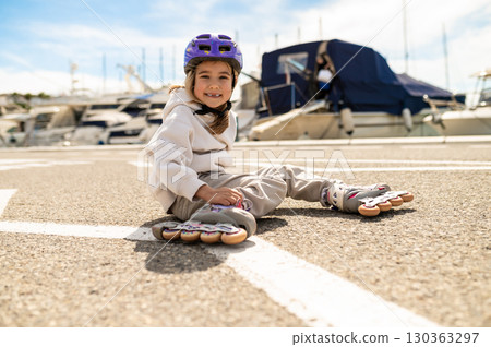 Smiling girl in hoodie and helmet sitting on rollerblades near yachts at sunny marina Smiling girl in hoodie and helmet sitting on rollerblades near yachts at sunny marina 130363297
