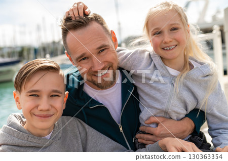 Father with two daughters smiling at camera on sunny marina pier Father with two daughters smiling at camera on sunny marina pier 130363354