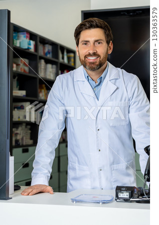 Dark-haired bearded pharmacist in a lab coat looking confident and contented Dark-haired bearded pharmacist in a lab coat looking confident and contented 130363579