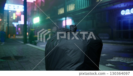 In a dimly lit urban environment, a large black trash bag sits on the sidewalk. Neon signs glow in the background, reflecting an almost eerie atmosphere during late night hours. 130364097