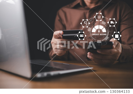With a hologram displaying a credit score report, a businessman uses a credit card and laptop to conduct financial analysis and manage online payments through advanced technology. 130364133